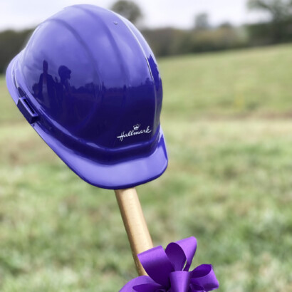 Purple hard hat resting on the handle of a gold shovel with a purple ribbon tied to it.